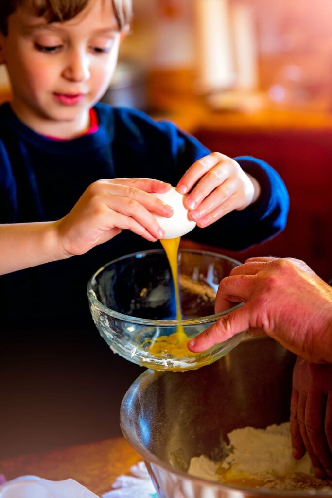 Young boy cracking egg into clear glass bowl.